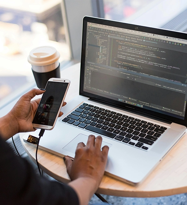 A person works on a laptop with code while holding a smartphone, a common scene for busy app developers.